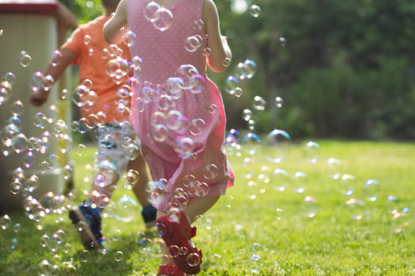 Kids are running in a grassy parkland area with a thick cloud of bubbles floating around them.