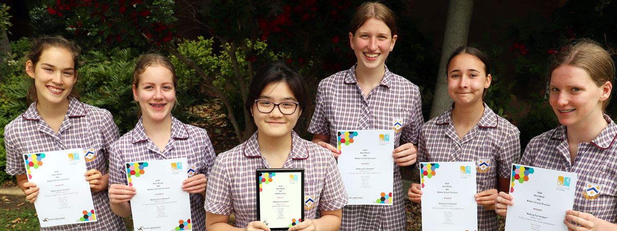 A group of girls in school uniform stand in a row, smiling widely; they are holding up their awards for participating in the Write a Book in a Day event.