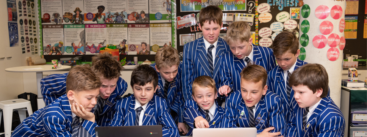 A group of boys wearing their school uniform blazers gather around a desk and 2 laptops; they are excitedly collaborating to write a book in a day.