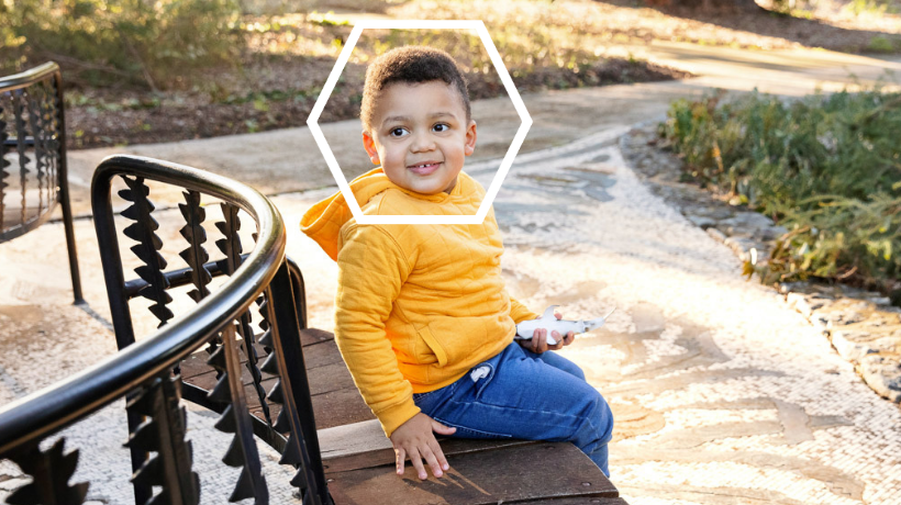 CJ smiles for the camera while sitting on a park bench; he is wearing a cheery, bright yellow hoodie