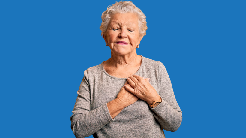 Coral stands peacefully in front of a blue backdrop with her eyes closed, a gentle smile on her face and her hands over her heart.