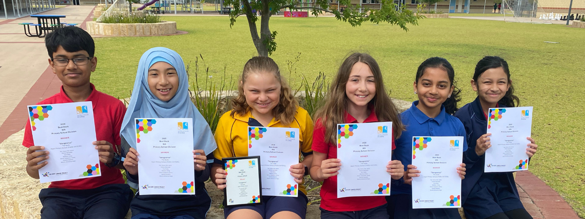 A group of younger students are very excited to be given their participation award certificates; they are smiling and sitting in a row along a bench in the school grounds.