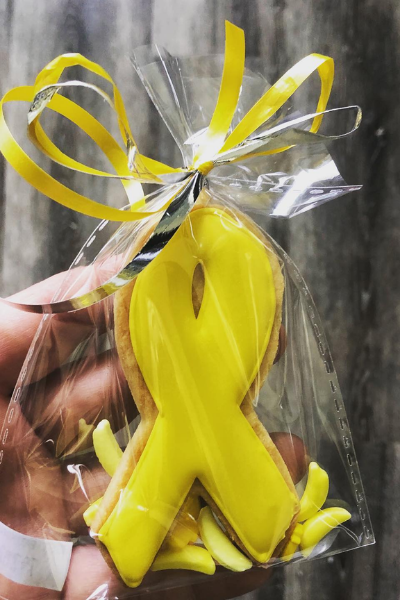 A tasty looking cookie in the shape of a childhood cancer ribbon is covered in bright golden-yellow icing and wrapped in a little cellophane bag, tied up with curly yellow and silver ribbons.