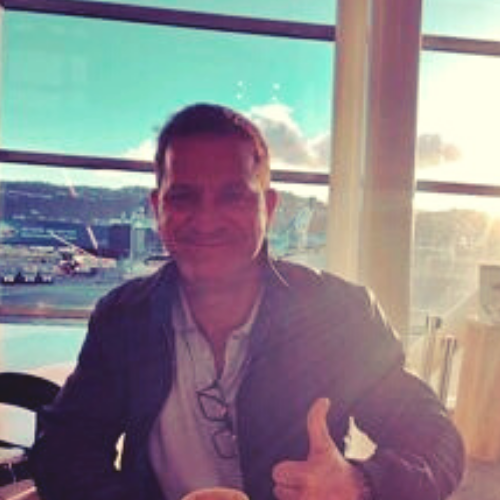 Craig McComb smiles and gives the camera a big thumbs up; he is sitting in an airport with the rising sun shining through the window behind him.