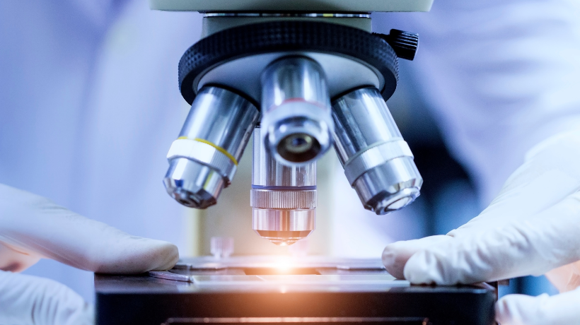 A close up image of a microscope; a researcher's hands are placing a slide under the lens and a bright light shines downwards to illuminate the sample being observed.