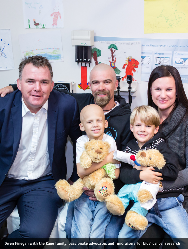 Owen and the Kane family smile for a photo in a clinic setting; Declan, undergoing cancer treatment, sits on his Dad Simon's lap while Declan's brother Brodie sits on Mum, Kim's lap.