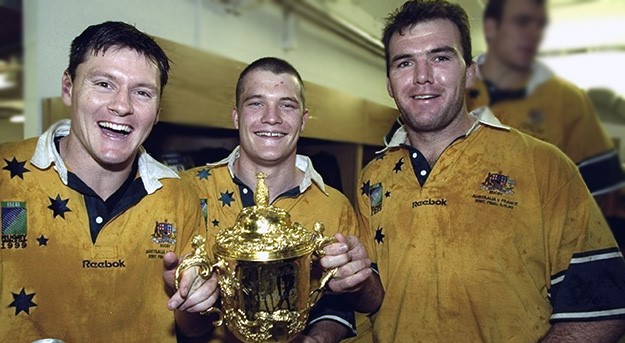 Owen Finegan and 2 teammates proudly hold their prize - a shiny gold cup - after the Wallabies' win at the 1999 Rugby World Cup.