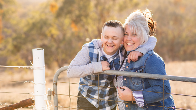 Alison Johansen and her son, Josh are smiling ear-to-ear and share a warm embrace while leaning on a gate at a rural property.