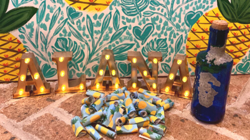 In front of a decorative backdrop featuring floral and heart patterns and large, bright pineapples sits a blue bottle sealed with a cork, the name 