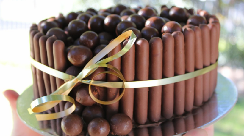 Atop a silver cake plate is a delicious looking circular cake; it is beautifully decorated - surrounded by chocolate covered finger biscuits placed vertically all around it and there are round chocolate balls (perhaps Malteasers) piled on top and cascading down one side of the cake; a gold ribbon tied into a bow has been tied around the circumference as a finishing touch. 