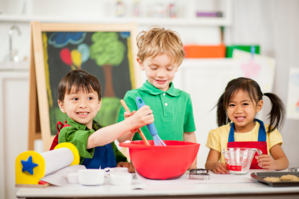 3 preschool aged children are excitedly gathered around red mixing bowl and measuring cups as they get ready to bake something for a good cause.