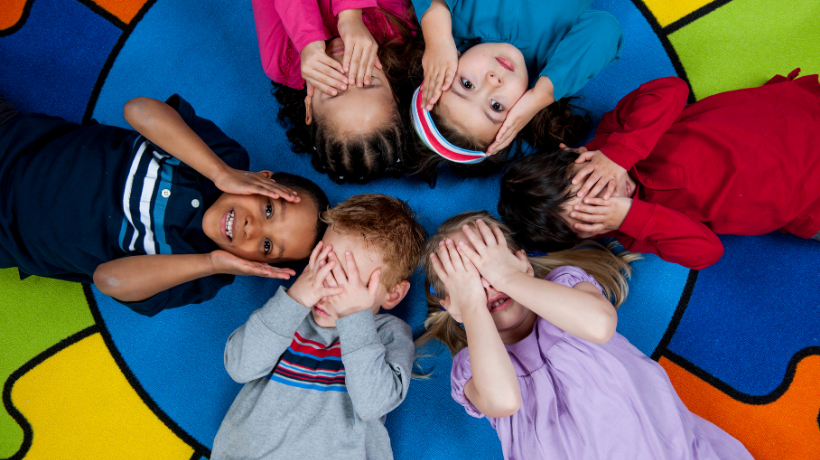 The image has been taken from above; Preschool aged children are lying on a brightly coloured mat that has a big blue circle in the centre - the children are positioned so that their heads meet in the middle of the circle.