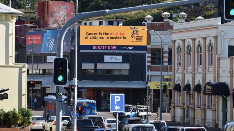 A busy urban intersection full of traffic and lined with buildings is pictured; a building in the centre of the image displays a large billboard advertising TKCP and raising awareness for childhood cancer.