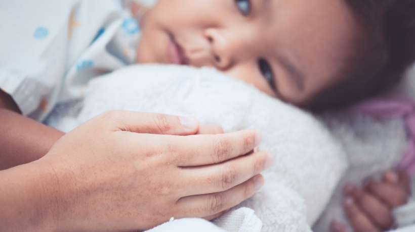 In the background, a small child wearing a hospital gown lies on their side looking up towards what we can assume is a parent sitting beside them out of frame; in the foreground, the parent grasps the child's little hand gently to provide some comfort.