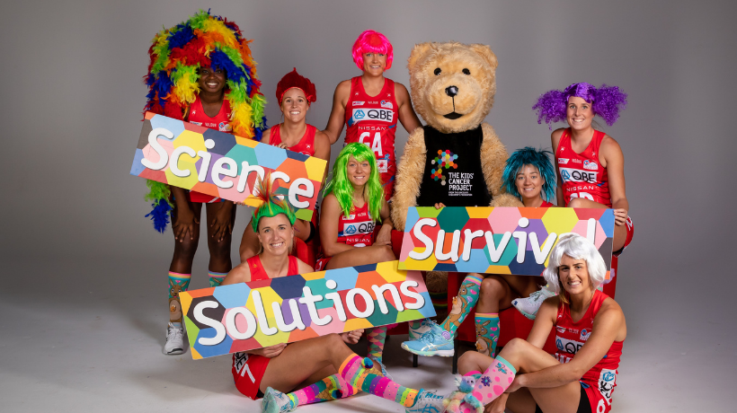 Netball players from the NSW Swifts gather for a group photo wearing their netball uniforms and some crazy, bright wigs and socks - with them stands the TKCP bear mascot; they are also holding 3 large posters covered with a colourful hexagon pattern and each one features a word in large white text: 