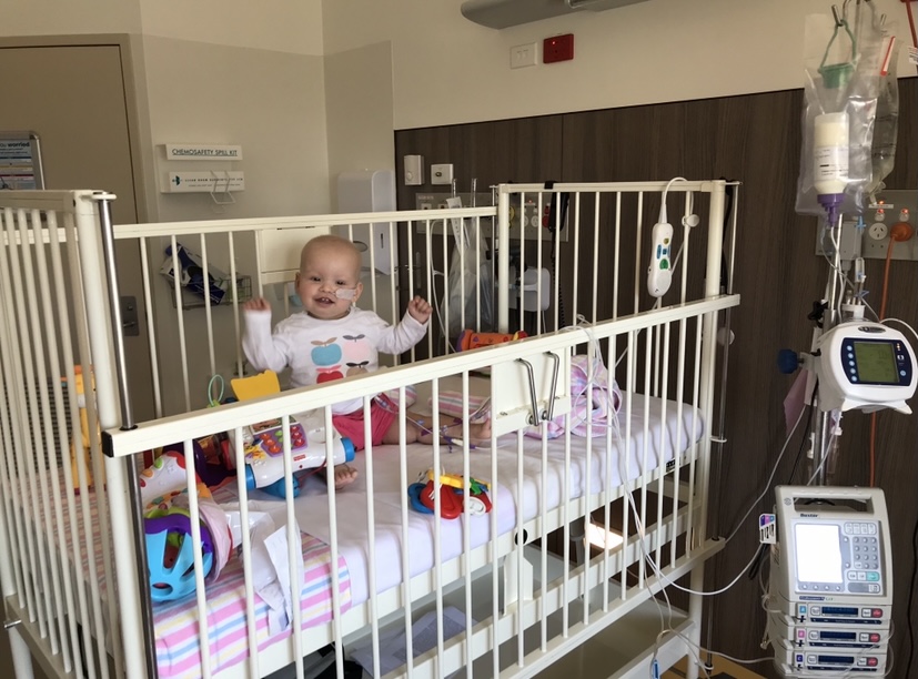 Adelaide sitting up in a cot filled with toys, medical equipment is to the right of the crib