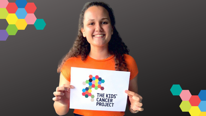 Anastassia smiles for the camera in front of a grey backdrop that is framed with a colourful hexagon pattern on opposing corners; she is holding an A4 size, white poster with the colourful TKCP logo printed on it.