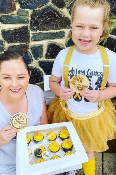 Millie and a baker from CJ's Sweets and Treats smile together while holding a box of delicious cupcakes with golden frosting and TKCP plaques.