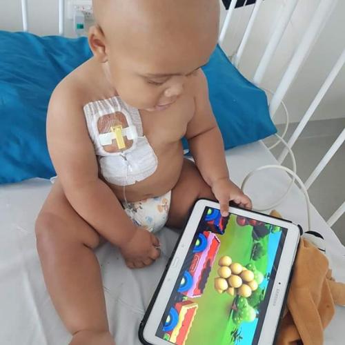 CJ sits in a hospital cot, watching a bright and fun cartoon on a tablet while he receives treatment via a port on his chest.