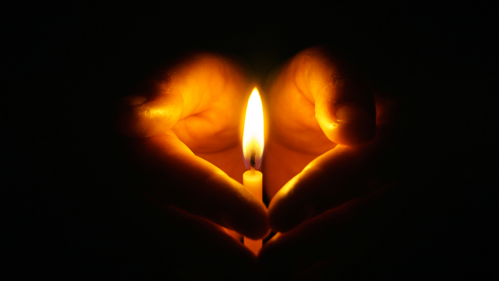 In the centre of a black background is a small candle, burning brightly - a pair of hands are cupped around it; the light from the flame illuminates the hands against the dark background, creating the illusion of a heart-shape.