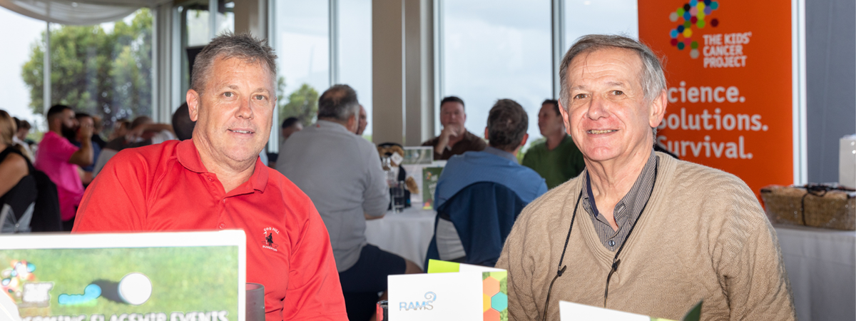 2 participants sit at their table for lunch and smile for the camera; behind them are other tables with participants engaged in lively conversation.