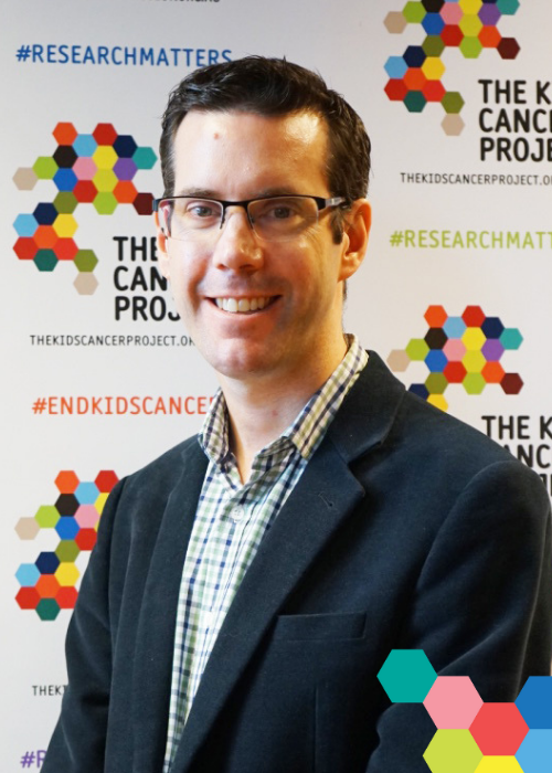 Professor Andrew Moore smiles while standing in front of a TKCP branded banner; a small cluster of colourful hexagons frames the image in the bottom-right corner.