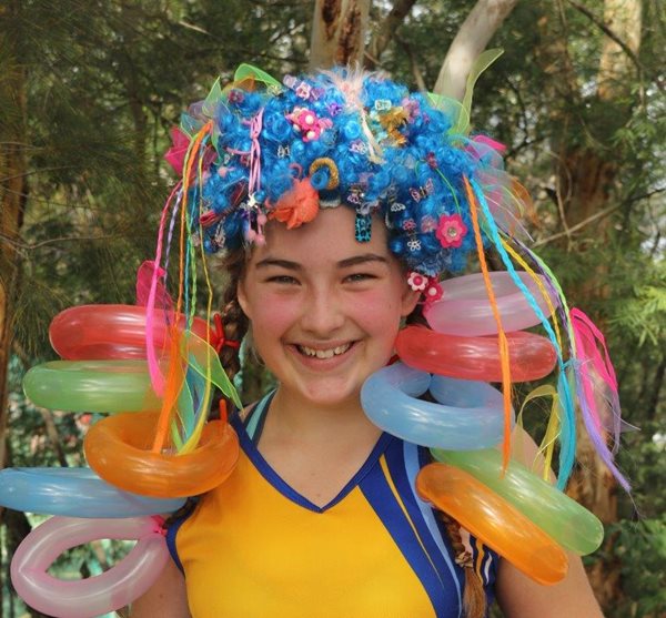 A student wears a short, blue, curly wig - it is covered in brightly coloured ribbons, clips and thin plaited strands of clip-in hair in crazy colours; some of these strands have balloons that have been tied into ring shapes attached to them.