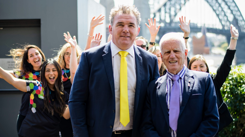 With the Sydney Harbour Bridge in the background, Owen Finegan and Col Reynolds smile for the camera; behind them is a happy group of people wearing TKCP tee-shirts.