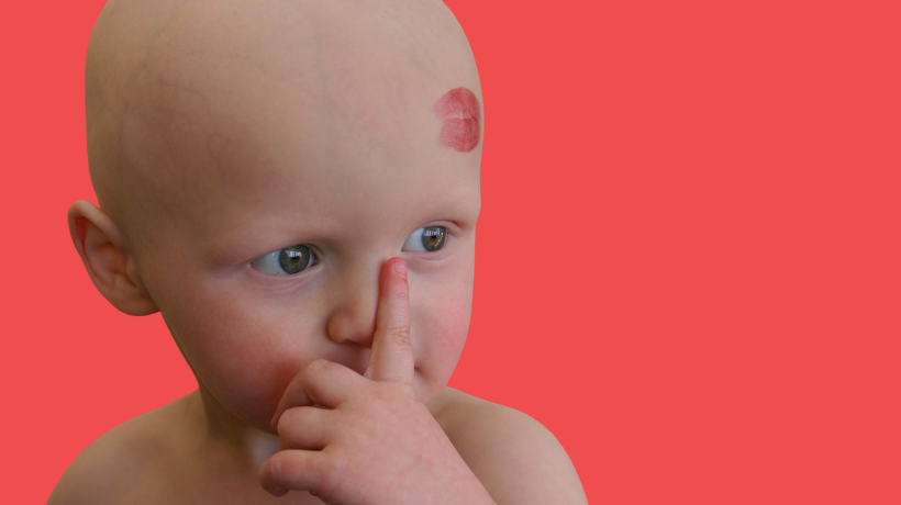 A small child who has lost their hair due to cancer treatment uses their index finger to touch their nose; on their forehead is a big red mark left by someone giving them a kiss and leaving lipstick behind. The backdrop is red.