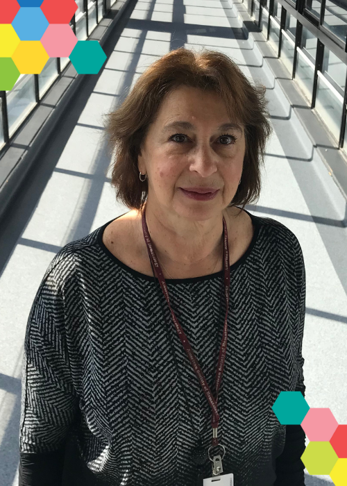 Dr Alla Dolnikov smiles while standing in the light-filled overpass of a research building; small clusters of colourful hexagons frame the image on opposing corners.