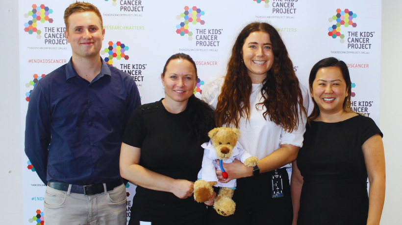 Members of the Colin Biggers & Paisley team smile for the camera holding a TKCP bear in a doctor costume; behind them is a banner featuring the TKCP logo and the hashtag #endkidscancer