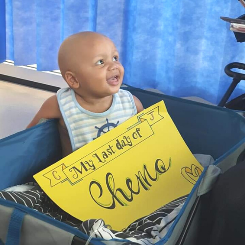 CJ smiles while sitting in the hospital cot; on his lap is a bright yellow poster that reads: 