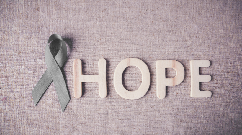 The image shows a light grey linen backdrop; placed thoughtfully atop the fabric is a silvery-grey brain cancer awareness ribbon followed by pale wooden letters that spell out the word 