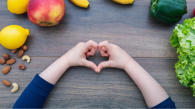 A birds-eye view of a wooden table; on the table is a tasty selection of healthy foods and snacks and in the centre of the image, we see a child's hands making a heart shape.