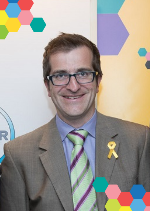 Dr Chris Frazer smiles in front of colourful banners at a kids' cancer event; small clusters of bright, colourful hexagons frame the image on opposing corners.