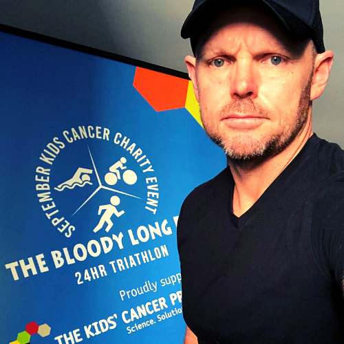 Mark Pacey, Cooper's Dad, stands in front of a promotional banner for The Bloody Long Day 24 hour triathlon.