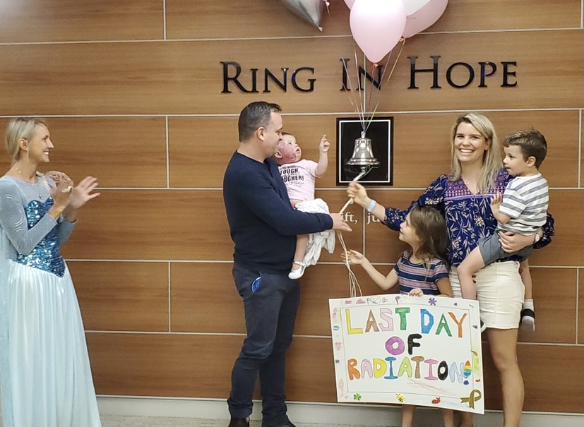 The family gathers around a bell with the words 'Ring In Hope' on a wall, along with pink balloons and a hand drawn sign reading 