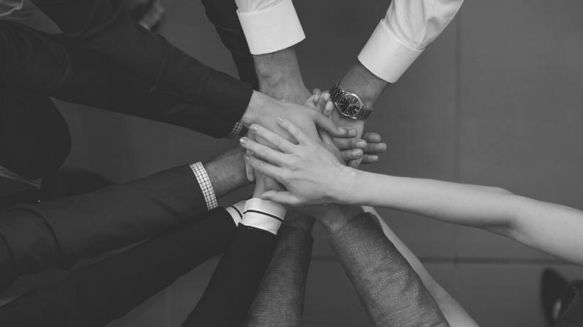 A black and white image depicts a close-up, birds-eye view of a group of people gathered in a circle, stacking their hands together in a team-huddle.