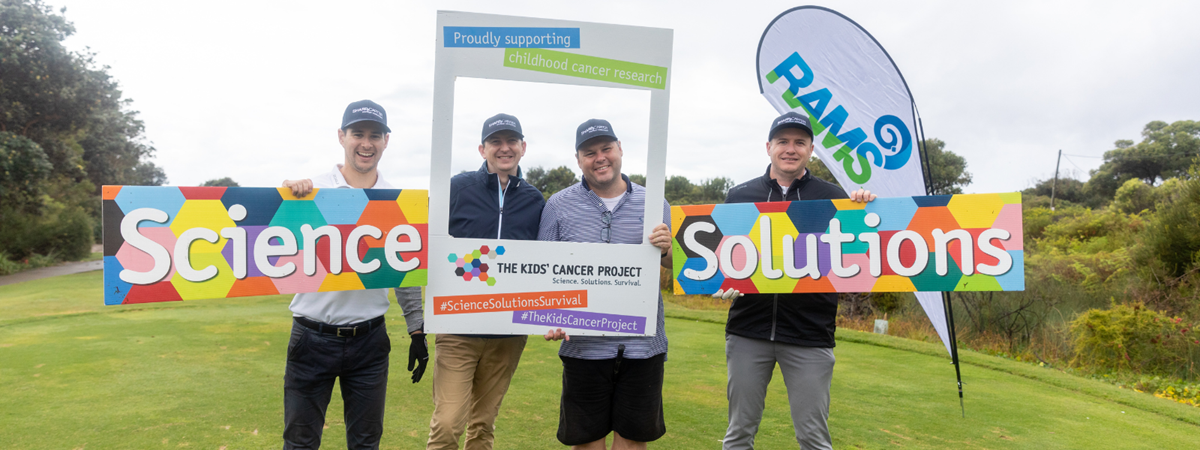 A golfing team poses for a photo holding up a frame featuring TKCP branding and the hashtags #ScienceSolutionsSurvival and #TheKidsCancerProject; they are also holing up banners decorated with brightly coloured hexagons that feature the words: "Science" and "Solutions"