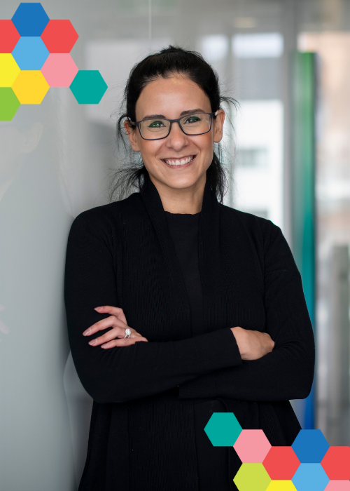 Dr Diane Hanna smiles broadly while leaning against the wall of a clinic hallway; a small cluster of brightly coloured hexagons frames the image on opposing corners.